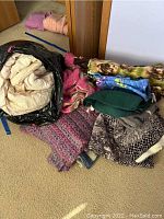 Pile of various linens including comforters, afghans, and throws stacked on floor with a black plastic bag containing some items.