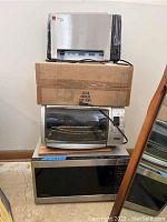 Three kitchen appliances stacked: Ronco Ready Grill on top with box, Oster toaster oven in middle with cable, Panasonic microwave oven at bottom