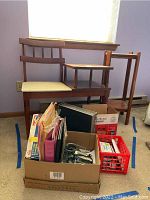Full view of vintage wooden telephone chair with attached table, with cream upholstered seat cushion, plant stand to the right, and boxes/crates of office supplies in front.