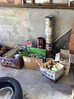 Overview photo of assorted used painting supplies including multiple paint cans and boxes with brushes and rollers in a garage setting.