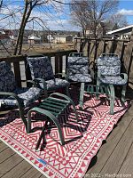 Four green metal patio chairs with blue and white floral cushions, two green metal ottomans with slatted webbing, red and white geometric outdoor rug beneath them.