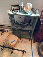 Wood painted green shelf with three shelves displaying metal decorative items including a spiral wine rack, iron holder, and hooks; beige woven basket with lid next to shelf, some scratches visible.