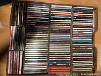 Photo of a box containing approximately 140 music CDs arranged flat, showing spines and fronts of jewel cases with various album titles and artist names.