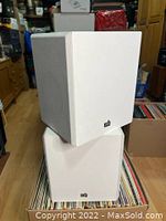 Front and angled view of two white PSB Ambient II dipole surround speakers stacked on vinyl records with white grills visible.