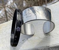 Two bracelets displayed; one wide silver-toned monogrammed cuff and one slimmer black ceramic bangle with floral design
