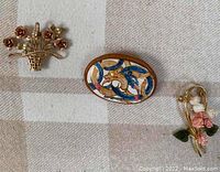 Three costume brooch pins, arranged on a checkered fabric background. Includes the basket flower pin, oval painted leather pin, and gold plated floral pin.