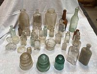 Photo showing a collection of 34 old bottles, two green glass insulators, and two clear glass cups arranged on a table.