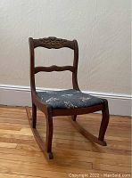 Full view of antique wooden rocking chair showing carved flower on upper back and blue fabric seat