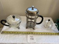 Set of three Bodum products including a large French press and two glass teapot infusers on a white cloth with measuring tape.