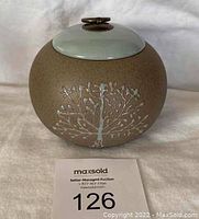 Front view of round ceramic tea jar with carved tree design and light blue lid with metal ring handle.