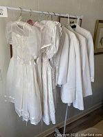 Photo of 4 girls white formal dresses and 4 boys white suits hanging on a rack, showing varying styles and fabric details.