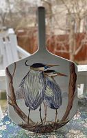 Front view of original painting of two great blue herons on a wooden pizza peel with a light lacquer finish and leather strap hanging.