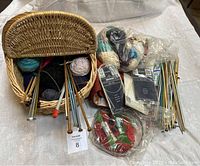 Wicker basket open showing yarn balls and knitting needles inside, plus additional yarn remnants, needles, and accessories next to basket.