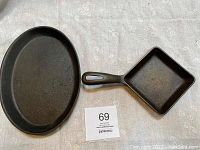 Oval Choice fajita pan and square Griswold egg skillet shown side by side on fabric surface, clearly showing the shape and handle design of each cast iron skillet.