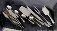 Top view of full flatware and serving utensil set laid on black fabric with pieces arranged horizontally including tongs, cutting tools, fork, spoons, and spatula.