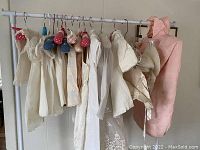 Wide view of various vintage baby clothes hanging on a rack displayed with several pieces in white and one pink garment at the side, multiple pairs of baby shoes also visible.