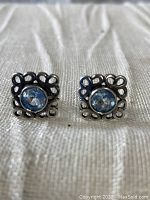 Close-up front view of the pair of blue topaz precision-cut faceted gemstones set in square 925 sterling silver pierced stud earrings with openwork decorative border. Fabric background.