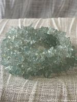 Stretch bracelet made of small, translucent aquamarine or dyed quartz tumble stones, shown coiled on a beige textile surface.