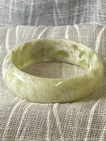 Photo of carved jade bangle showing side and front views, placed on a cushion with a textured linen background.