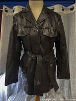 Full front image of the chocolate brown leather coat on mannequin, showing lapel collar, button closures, tie belt, and four pockets.