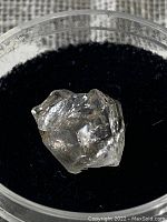 Raw, uncut diamond in a transparent container on black fabric background showing facets and natural crystal form.
