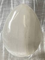 Polished white selenite egg with natural internal striations against a fabric backdrop.