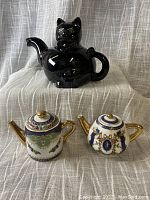 Photo showing all three teapots: one black ceramic cat-shaped teapot and two smaller Adeline fine porcelain teapots with decorative gold and blue detailing.