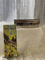 Full view of Echo Harp harmonica next to original decorative box with mountain cabin artwork.