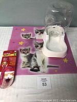 Photo showing white automatic water dispenser with clear water container placed on a purple cat feeding placemat with kitten images and star designs, and kitty toothbrush in packaging beside it.