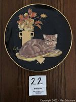 Front view of a round porcelain plate with a cat lying on a cushion beside a vase of flowers on a black background with gold rim, signed by Aldo Fabio.
