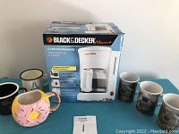 Photo showing boxed Black & Decker 5-cup coffee maker with five coffee mugs surrounding it: including a pink donut-shaped mug, two black mugs, and two mugs with outdoor or nature designs.