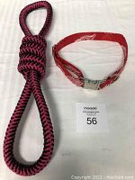 Photo showing the pink and black braided tug-of-war rope alongside the red Detroit Red Wings dog collar with metal buckle clasp lying on a white surface.