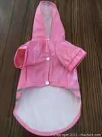 Pink shiny dog raincoat with hood displayed on wooden surface, front view, showing snap buttons and short sleeves.