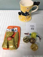 Photo of hand painted yellow ceramic mug with black cat, small black cat figurine on handle, yellow floral background, matching saucer with floral design and small black cat figurine, plus set of miscellaneous cat toys including gold mesh-covered catnip toys, green ball with ropes, and jingle bells.