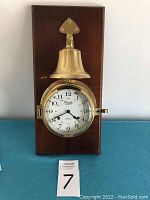 Full view of the ship clock mounted on a rectangular wood plaque with a brass bell above and white clock face below.