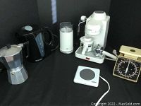 Photo showing all coffee appliances including espresso maker, grinder, kettle, warmer, percolator, scale, and timer arranged on black background.