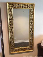 Full view of the large rectangular ornate wall mirror hanging on a wall, showing gold gilt carved frame with floral and geometric patterns around the clear mirror glass.