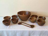 Full set of one large teak bowl, eight smaller bowls and two serving utensils on a white surface against plain wall.