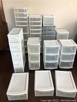 Full view of 12 assorted plastic storage drawer units in varying sizes and drawer counts, arranged on a hardwood floor next to a wall and furniture.