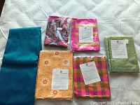 Five pieces of folded Thai silk fabric in different colors and patterns laid on a white sheet, showing variety of silk textiles.