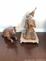 Full view of Thai statue on wooden base and reclining elephant candle holder side by side.