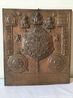 Front view of copper plaque showing central mandala, Tibetan script, symbolic calendar circles, and three raised deity figures at top center.