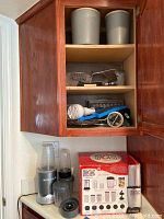 Open cabinet shelf with two grey dry goods canisters on top shelf, metal graters and utensils on middle shelf, handheld mixer and other utensils on bottom shelf. Below cabinet is Magic Bullet blender set in box and NutriBullet blender with blending cups on counter.