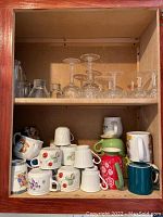 Shelves displaying assorted ceramic mugs with various designs and holiday themes, several clear glass stemware including wine glasses, and a glass carafe.
