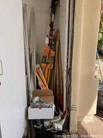 Photo showing an assortment of long handled garden tools including pole saw, shovel, and other tools along a wall corner