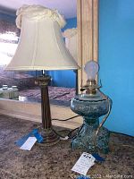 Photo showing both lamps side by side on a countertop with a mirror behind them. The wooden lamp has a cream-colored fabric shade with bows, the vintage glass base lamp has no shade and a visible bulb.