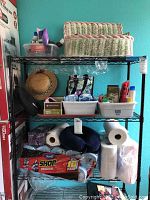 Overall view of metal rack loaded with assorted paper towels, wipes, and sanitary products including a woven basket on top shelf and a blue neck pillow on middle shelf.