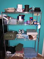 A metal shelving unit stocked with various household items including folded linens, electric blood pressure monitor, food storage containers, paper products, and small miscellaneous items like candles and ceramic pot.
