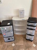 Four plastic storage units: three black or white framed units with three or four clear drawers standing against the wall and one mini unit with five drawers on top of the white unit.