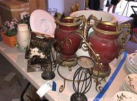 Photo showing two large red and gold decorative vases, brass candlestick holders, faux flowers, plant pot, and other items on a table.